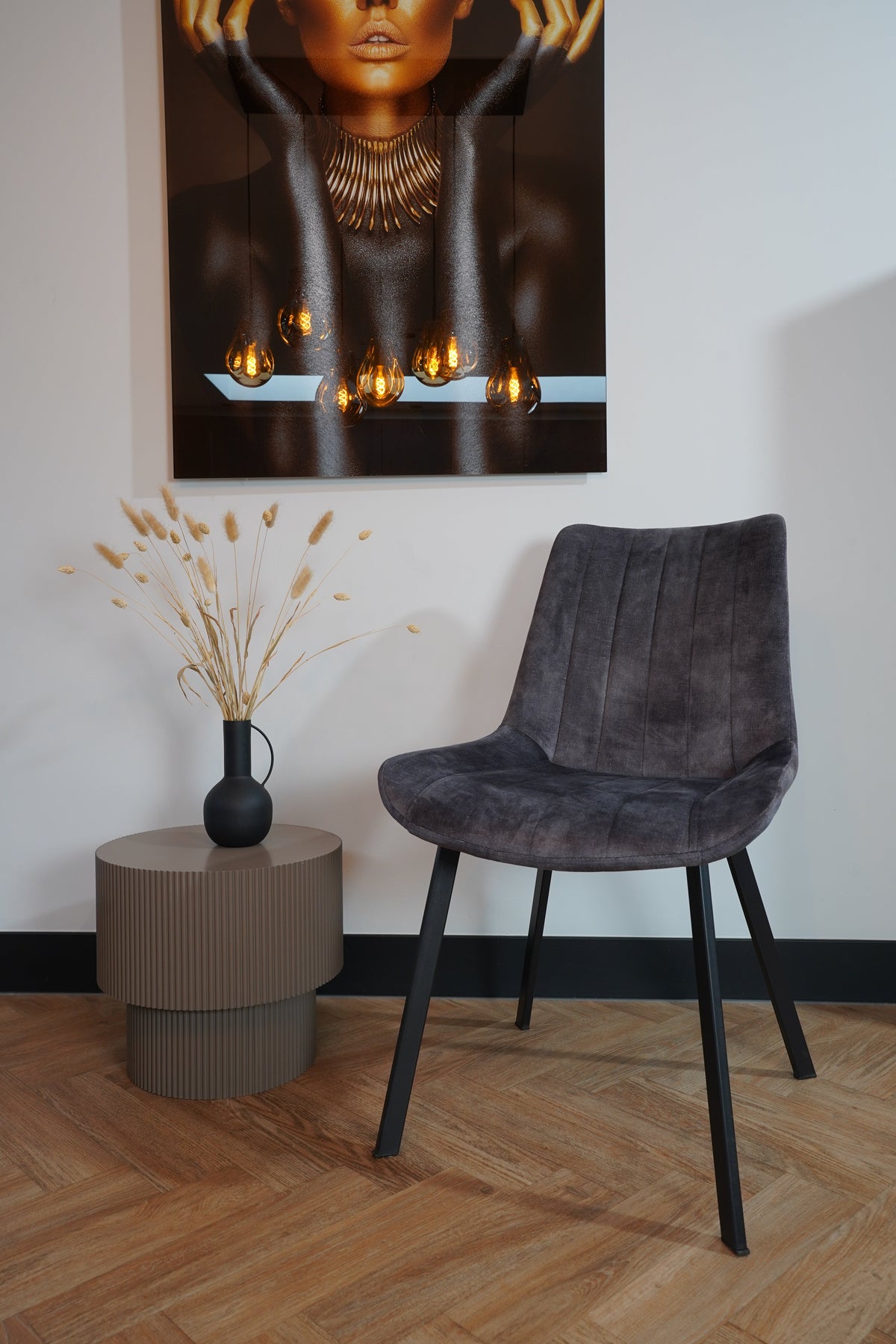 Eetkamerstoel Marilène in grijze velvet, moderne poten, naast bijzettafel met vaas en kunstwerk aan de muur.